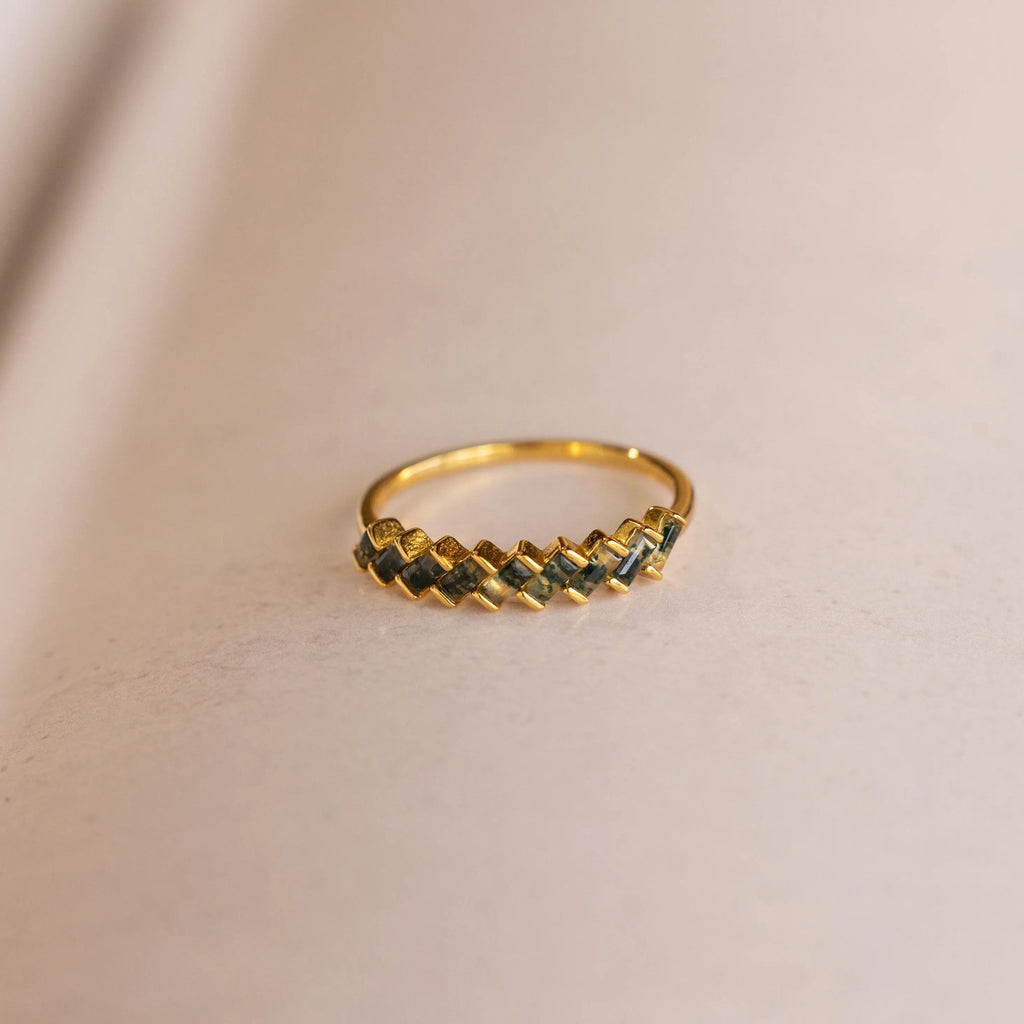 The Moss Agate Eternity Ring features a zigzag chevron-patterned gold band, inspired by nature and beautifully displayed on a light beige surface—an elegant choice for any occasion.
