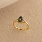 The Agate Teardrop Ring in 18K Gold features a pear-shaped green gemstone and small diamonds, set on a beige textured background—reminiscent of a vintage cocktail ring and perfect for making a statement.