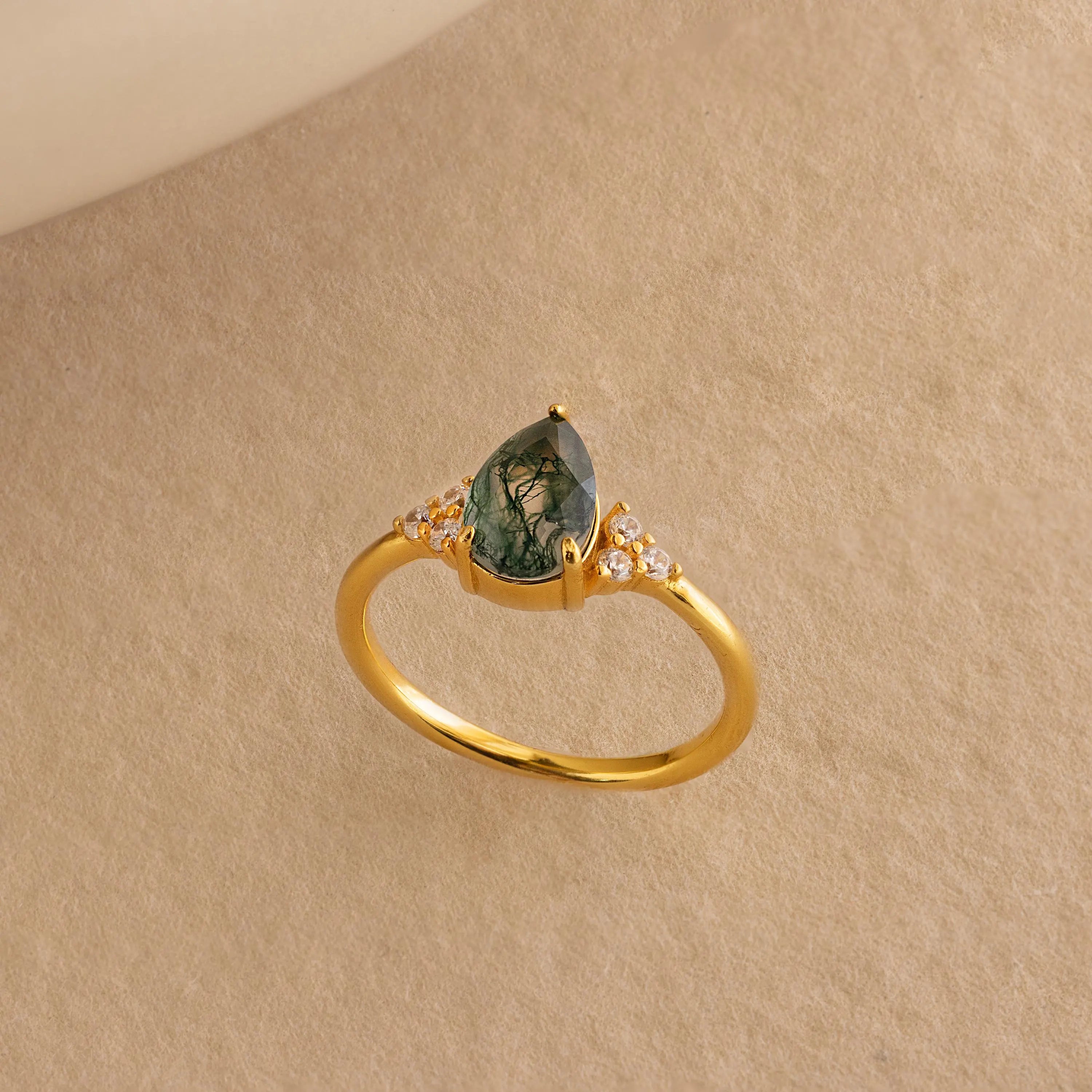 The Agate Teardrop Ring in 18K Gold features a pear-shaped green gemstone and small diamonds, set on a beige textured background—reminiscent of a vintage cocktail ring and perfect for making a statement.