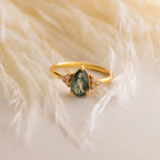 The Agate Teardrop Ring features a gold band with a green teardrop gemstone and clear stones, displayed on a cream surface. This elegant ring is a perfect cocktail accessory for any occasion.