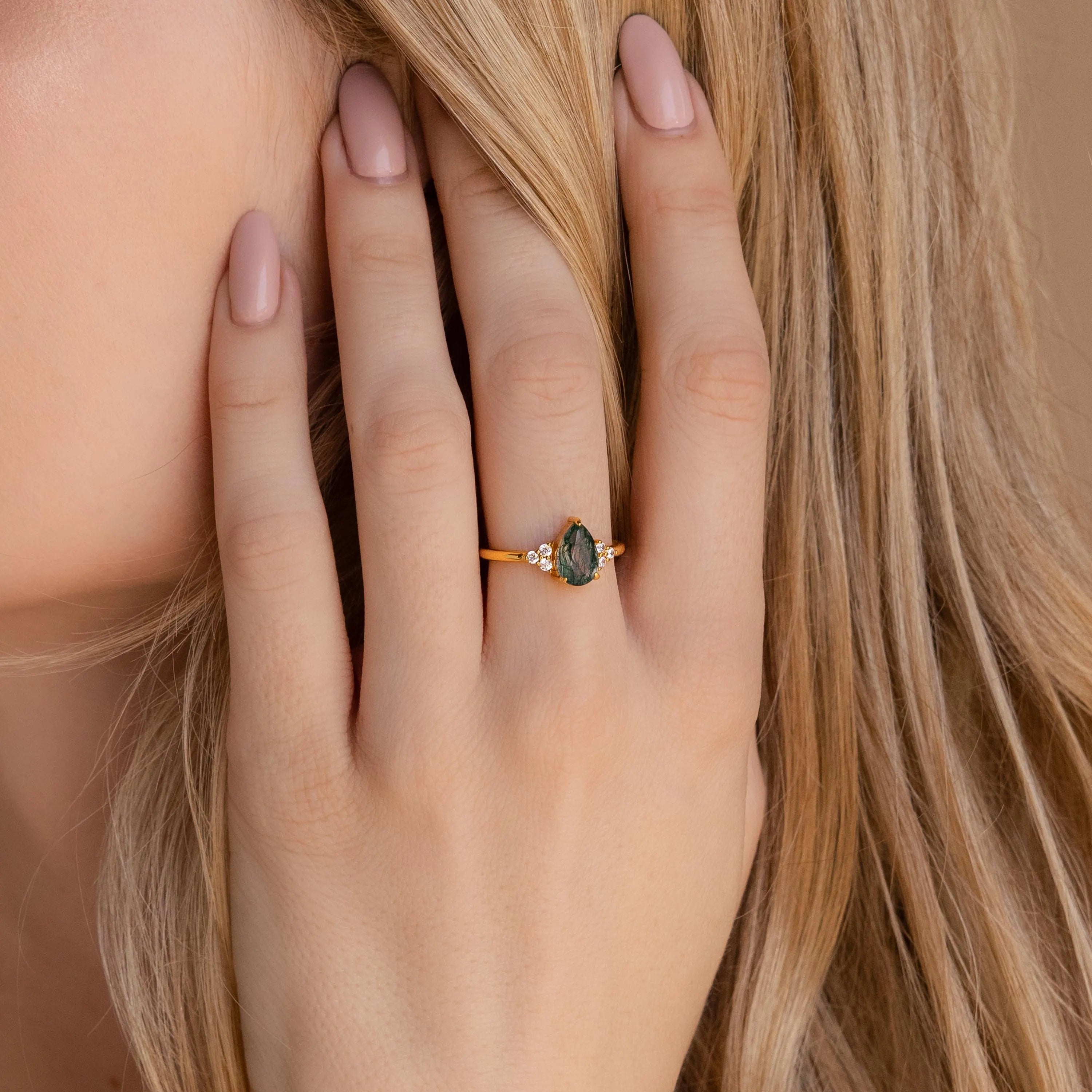 A hand with nude nails wears the Agate Teardrop Ring, showcasing a green moss agate gemstone and sparkling accent stones set in gold.