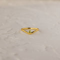 The Peridot Drop Curve Ring—a delicate gold stacking ring featuring a small green peridot gemstone—rests on a textured white surface.