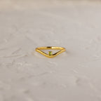 The Peridot Drop Curve Ring—a delicate gold stacking ring featuring a small green peridot gemstone—rests on a textured white surface.