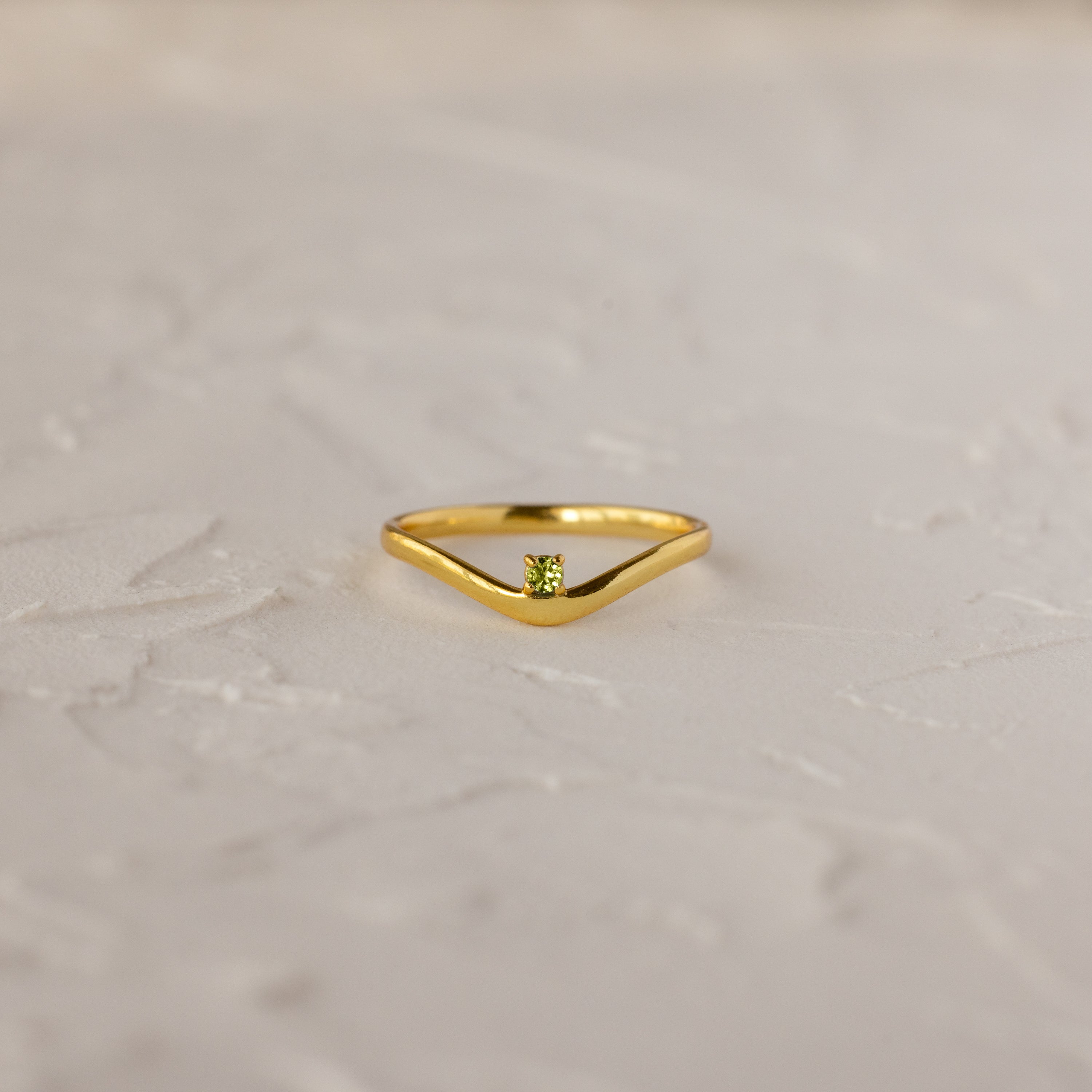 The Peridot Drop Curve Ring—a delicate gold stacking ring featuring a small green peridot gemstone—rests on a textured white surface.