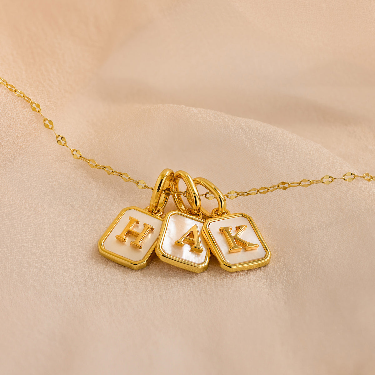 A gold chain necklace with three square pendant charms featuring white pearl inlay and raised gold letters 'H', 'A', and 'K', displayed on a soft beige fabric background.