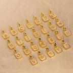 Gold-plated square letter charms spelling out the alphabet, each with a white inlay and a small lobster clasp at the top, arranged on a textured beige surface.