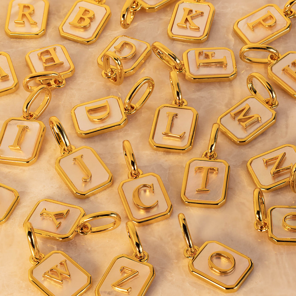 A close-up arrangement of gold letter charms with white pearl enamel backgrounds, each featuring a raised uppercase letter and a small jump ring, photographed on a light neutral surface.