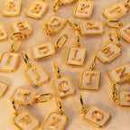A close-up arrangement of gold letter charms with white pearl enamel backgrounds, each featuring a raised uppercase letter and a small jump ring, photographed on a light neutral surface.
