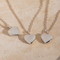 Three silver heart-shaped locket necklaces on delicate chains, displayed on a textured beige stone surface. The center pendant is engraved with a monogram letter.