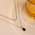 A silver-toned double-layer necklace with a black oval pendant, styled on a beige textured surface next to a glass of amber-colored liquid.