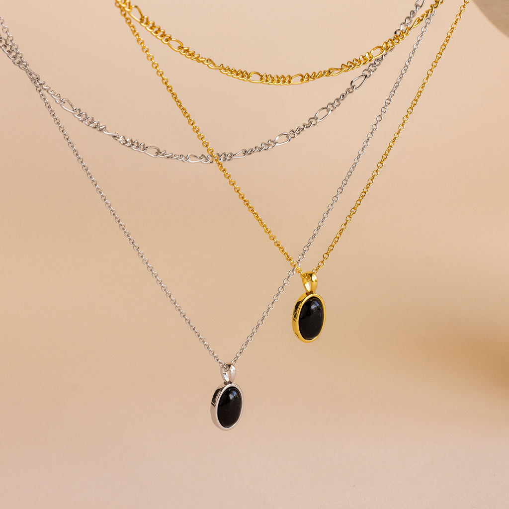 Three layered necklaces displayed on a neutral beige background: a silver chain with a black oval pendant, a gold chain with a black oval pendant, and a silver Figaro chain with a black oval pendant.