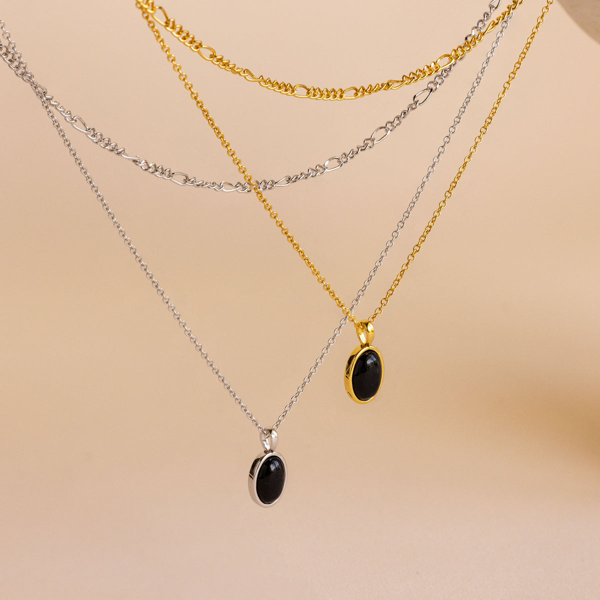 Three layered necklaces displayed on a neutral beige background: a silver chain with a black oval pendant, a gold chain with a black oval pendant, and a silver Figaro chain with a black oval pendant.