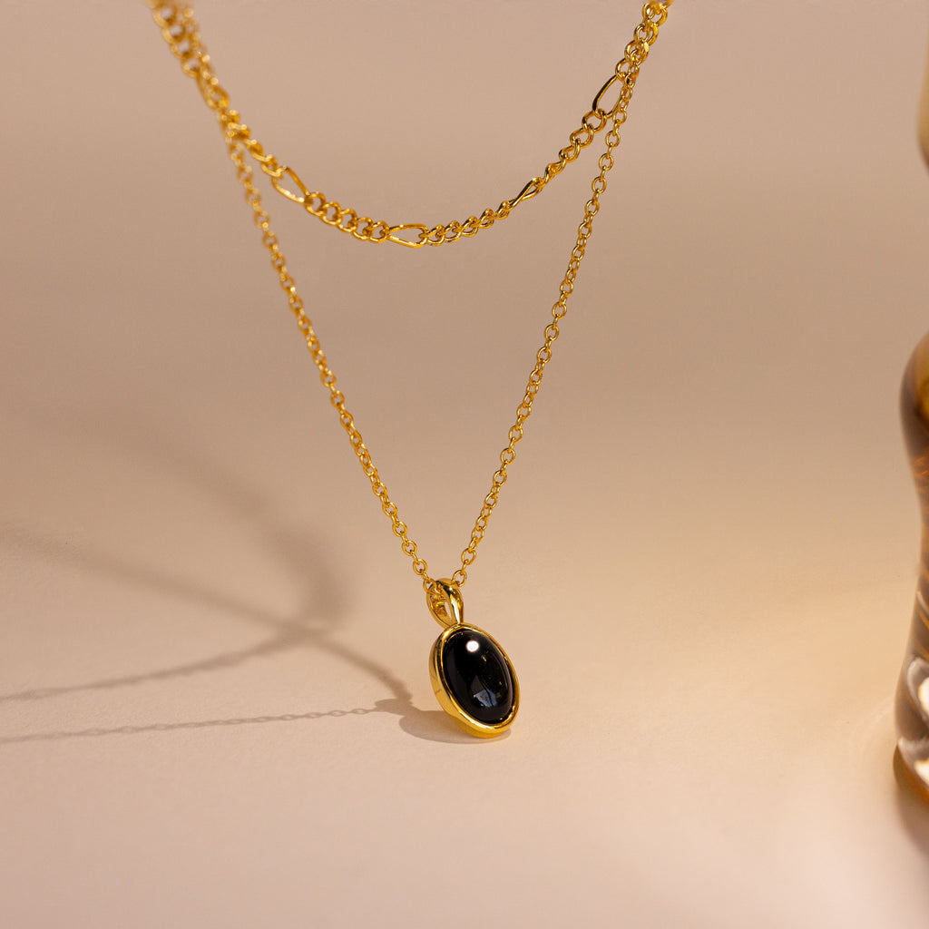 A gold layered necklace featuring a delicate chain and a longer chain with an oval black stone pendant, displayed on a warm beige surface.