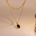 A gold layered necklace featuring a delicate chain and a longer chain with an oval black stone pendant, displayed on a warm beige surface.