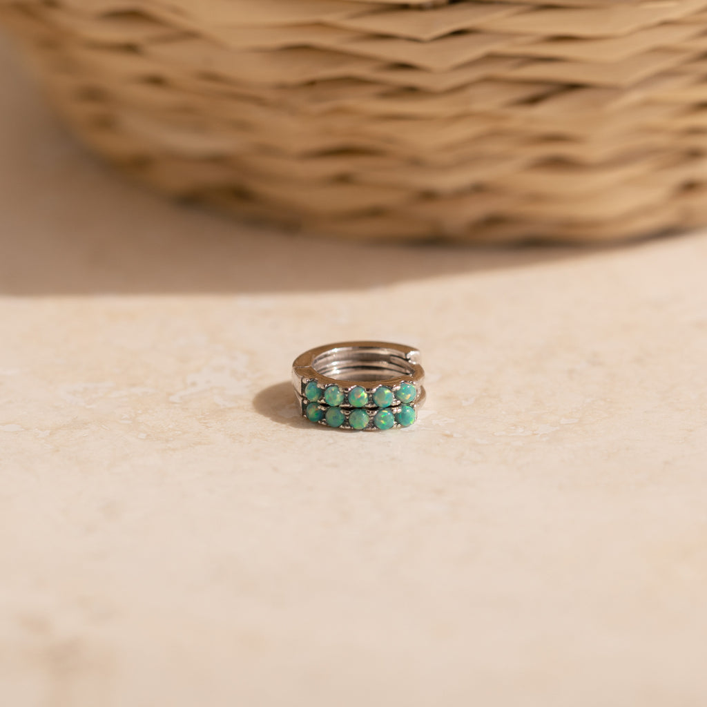 Our Garden Green Opal Huggies in silver resting on a light beige stone surface with a blurred woven basket in the background.