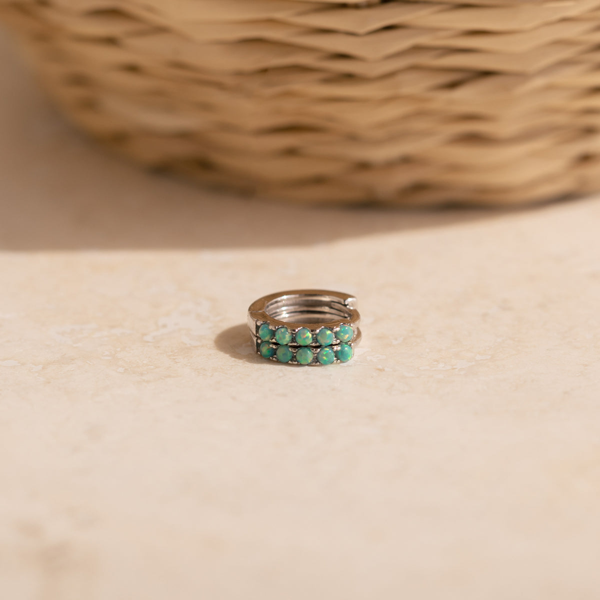 Our Garden Green Opal Huggies in silver resting on a light beige stone surface with a blurred woven basket in the background.