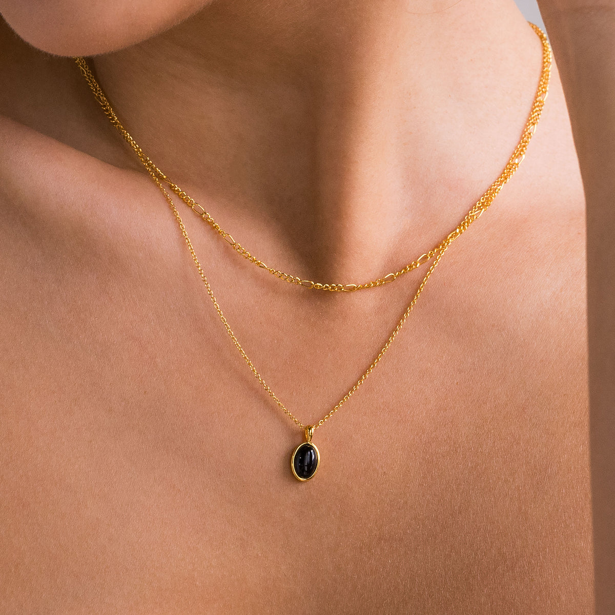 A close-up of a model's neck and upper chest wearing a delicate gold double-layer necklace with a small oval black pendant.