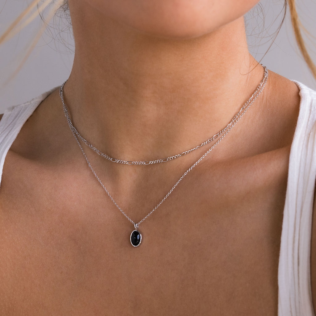 A close-up of a model wearing a double-layered silver necklace. The top layer features a delicate curb chain, and the bottom layer features a fine link chain with an oval black pendant set in a silver bezel.