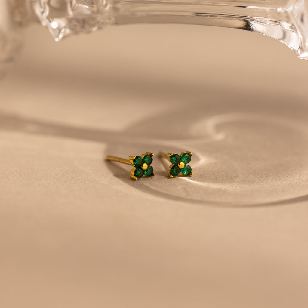 A pair of small gold stud earrings with four green gemstones arranged in a flower shape, resting on a warm beige surface with a soft shadow and part of a clear glass object at the top.