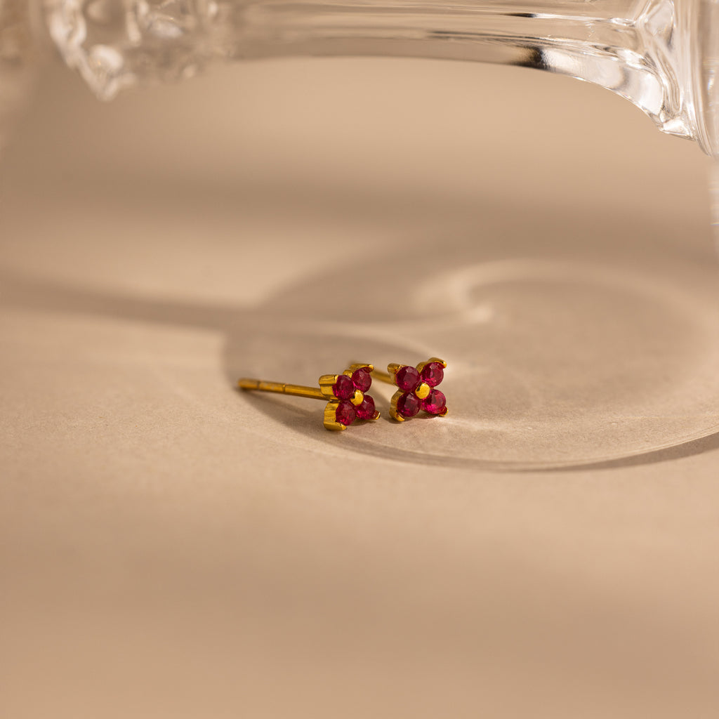 A pair of small gold stud earrings with four pink ruby gemstones arranged in a flower-like cluster, photographed on a warm beige surface with a clear glass object partially visible at the top.