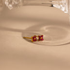 A pair of small gold stud earrings with four pink ruby gemstones arranged in a flower-like cluster, photographed on a warm beige surface with a clear glass object partially visible at the top.