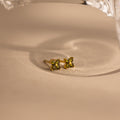 A pair of small gold stud earrings with four green gemstones arranged in a flower shape, resting on a beige surface with a soft shadow.