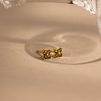 A pair of small gold stud earrings with four green gemstones arranged in a flower shape, resting on a beige surface with a soft shadow.