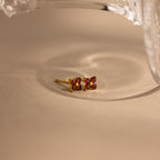 A pair of gold-toned stud earrings with four small red gemstones arranged in a square, resting on a beige surface with a soft shadow and a glass object partially visible at the top.