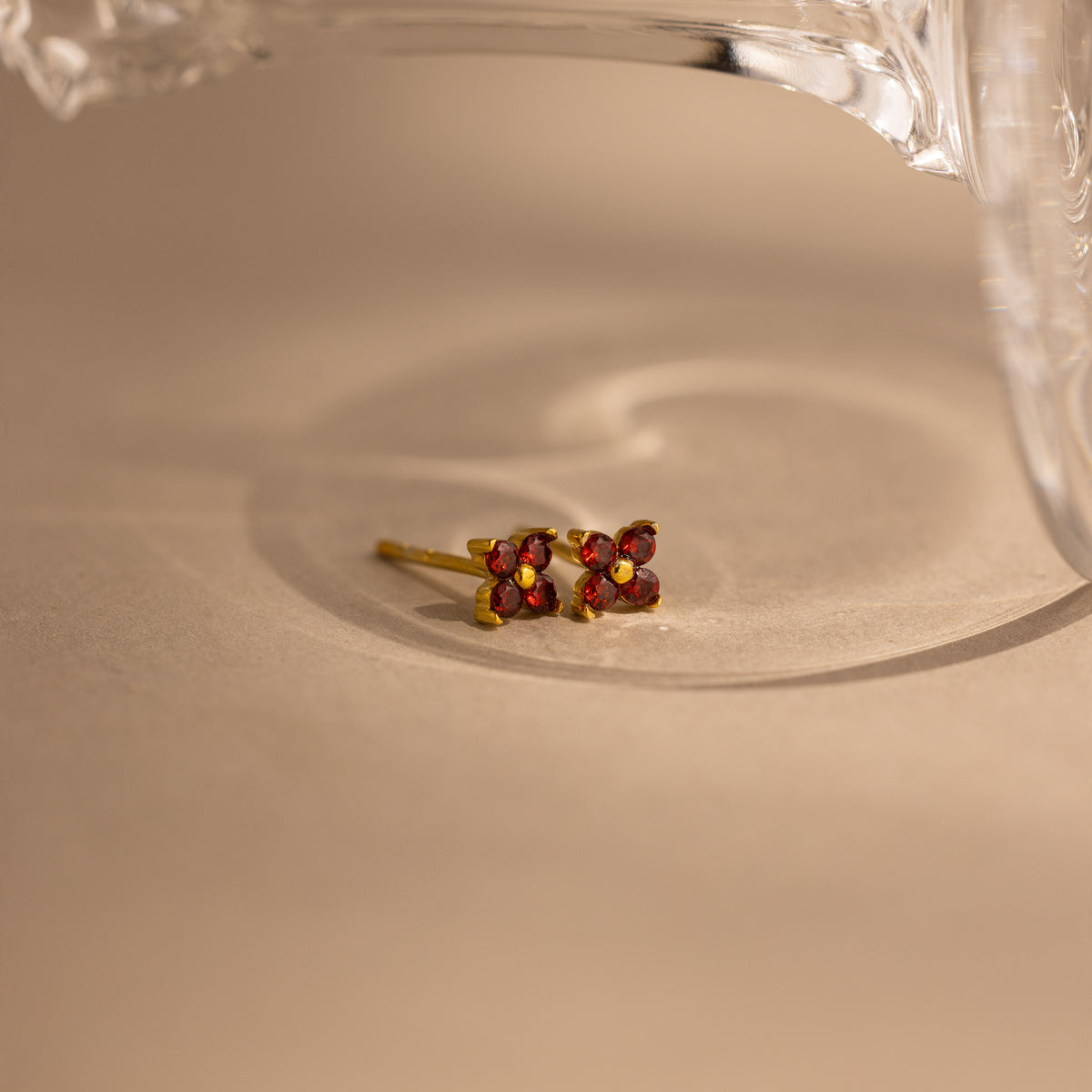 A pair of gold-toned stud earrings with four small red gemstones arranged in a square, resting on a beige surface with a soft shadow and a glass object partially visible at the top.