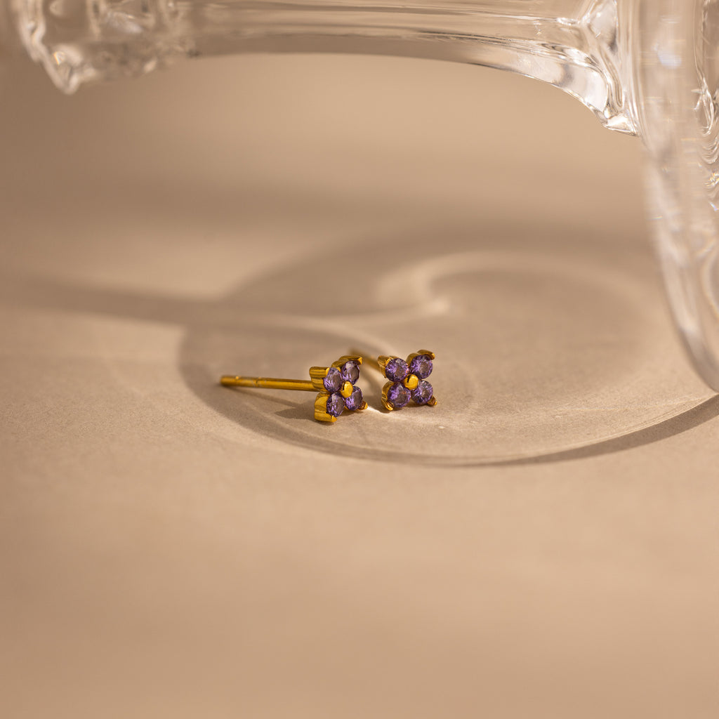 A pair of small gold stud earrings with four purple gemstones arranged in a flower-like cluster, photographed on a warm beige surface with a clear glass object partially visible at the top.