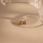 A pair of small gold stud earrings with four purple gemstones arranged in a flower-like cluster, photographed on a warm beige surface with a clear glass object partially visible at the top.