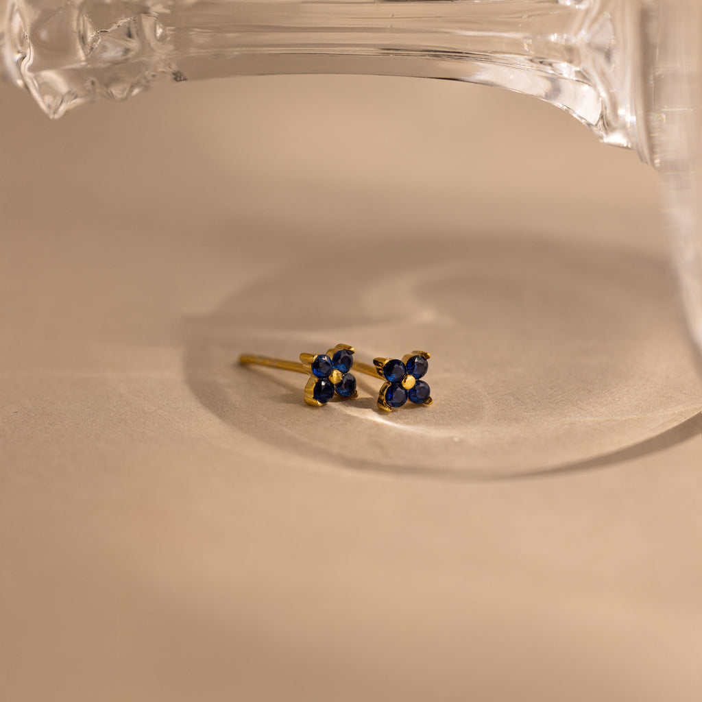 A pair of small gold stud earrings with deep blue flower-like faceted stones, resting on a beige surface under a clear glass object.