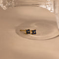 A pair of small gold stud earrings with deep blue flower-like faceted stones, resting on a beige surface under a clear glass object.