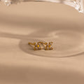 A pair of small gold stud earrings with four clear stones arranged in a flower shape, resting on a beige surface with a soft shadow and a blurred clear object at the top.