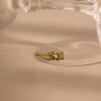 A pair of small gold stud earrings with four light blue stones, resting on a beige surface with a soft-focus glass object in the background.