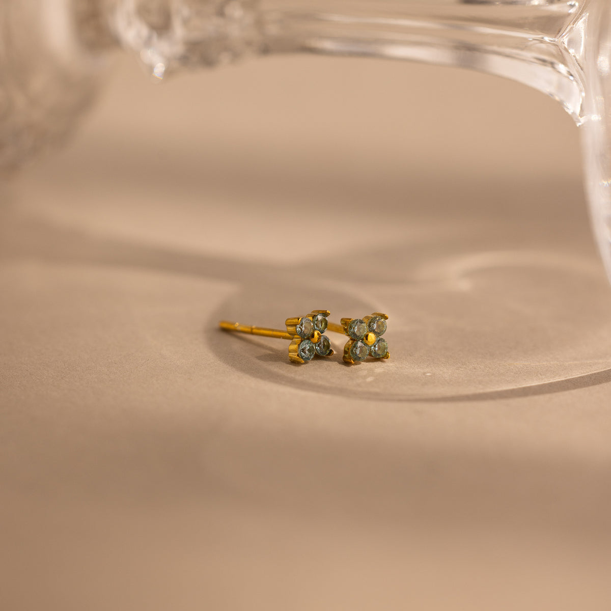 A pair of small gold stud earrings with four light blue stones, resting on a beige surface with a soft-focus glass object in the background.