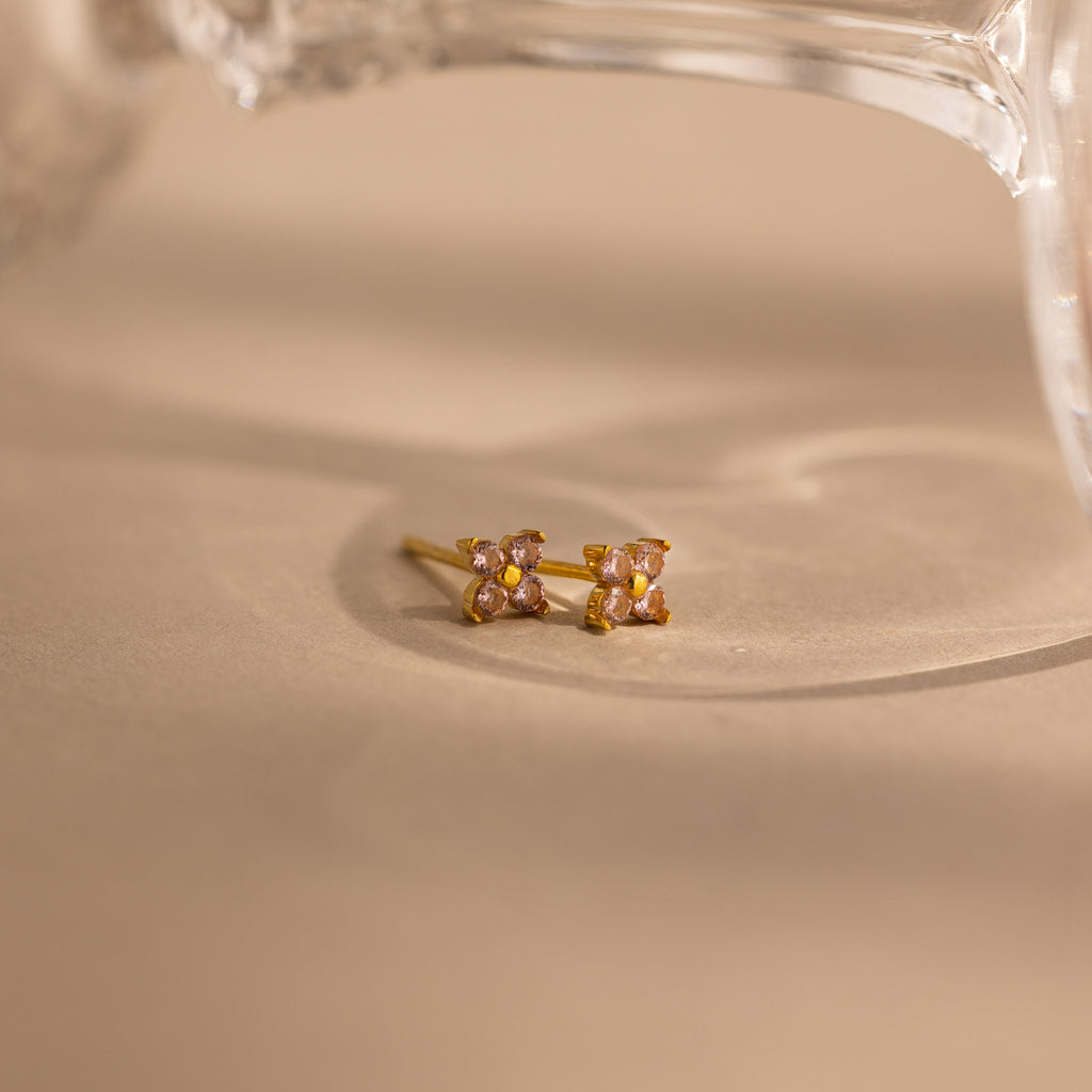 A pair of small gold stud earrings with four light pink stones arranged in a square, resting on a beige surface with a soft-focus glass object in the background.