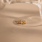 A pair of small gold stud earrings with four light pink stones arranged in a square, resting on a beige surface with a soft-focus glass object in the background.