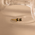 A pair of small gold stud earrings with black flower-like settings, photographed on a warm beige surface with a blurred clear glass object in the background.