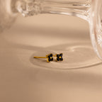 A pair of small gold stud earrings with black flower-like settings, photographed on a warm beige surface with a blurred clear glass object in the background.