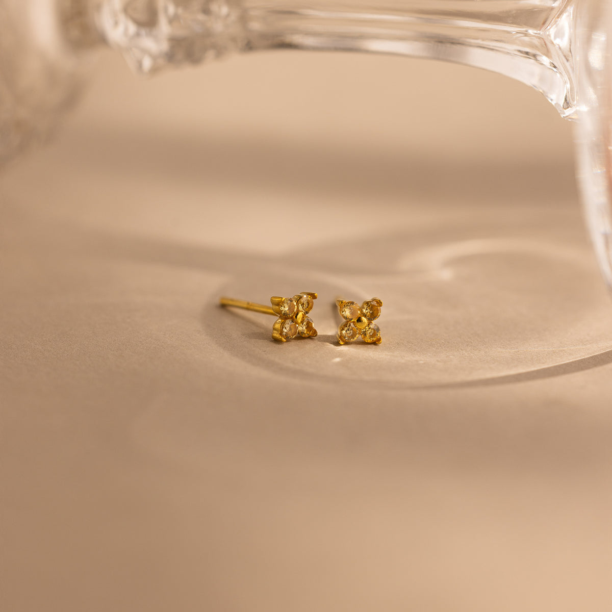 A pair of small gold stud earrings with a butterfly-like design, resting on a beige surface with a soft-focus glass object in the background.