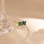 A pair of small gold stud earrings with four blue green gemstones arranged in a flower shape, resting on the clear glass base of a wine glass against a warm beige background.