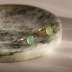 Minimalist oval green gemstone rings in gold and silver finishes, featuring smooth pastel green stones in delicate bezel settings, displayed together on a marble jewelry tray.