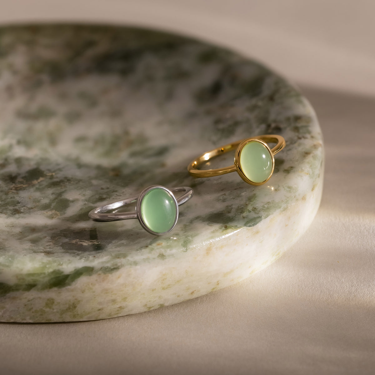 Minimalist oval green gemstone rings in gold and silver finishes, featuring smooth pastel green stones in delicate bezel settings, displayed together on a marble jewelry tray.