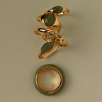 Five gold rings with green stones, including the Haven Jade Spinner Ring, are stacked above a round green and gold band bracelet on a beige background—an elegant display of mindful jewelry.