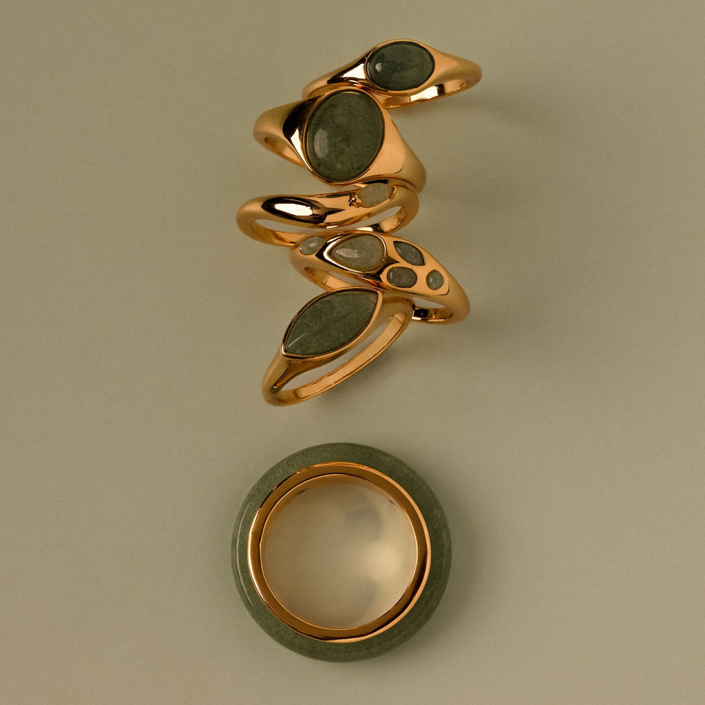 Five gold rings with green jade stones, stacked above a Jade Dewdrop Ring bracelet, radiate natural elegance on a beige background.