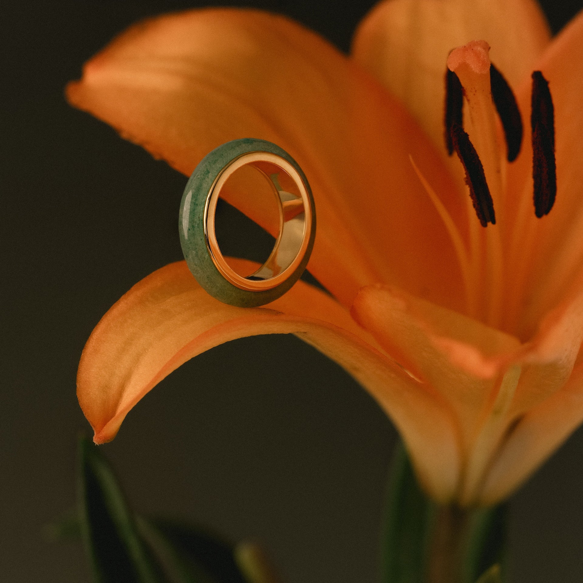 The Haven Jade Spinner Ring rests delicately on the petal of an orange lily, its gold band shimmering against a dark background.