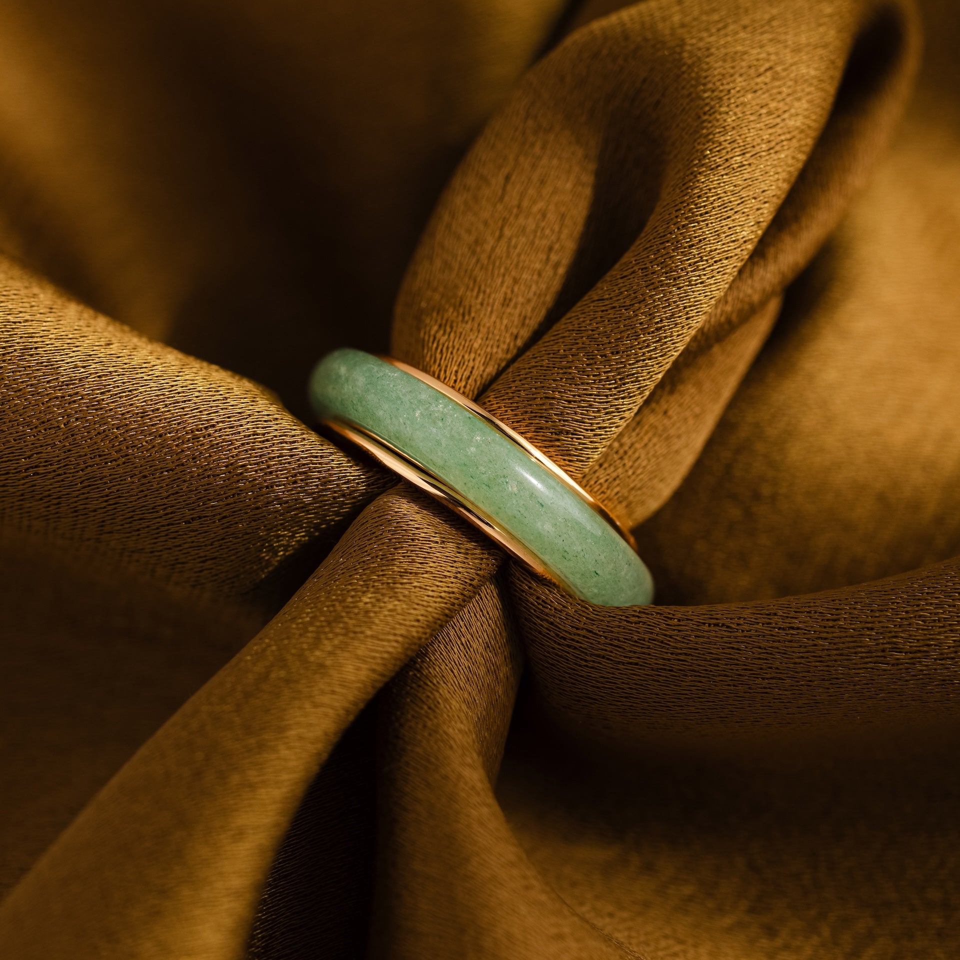 The Haven Jade Spinner Ring features a green jade band and gold accents, displayed on shimmering brown fabric—an elegant and mindful jewelry piece.