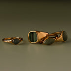 Four gold rings with green and gray stone inlays, including the Malachite Signet Ring—featuring malachite, the stone of transformation—displayed on a neutral surface.