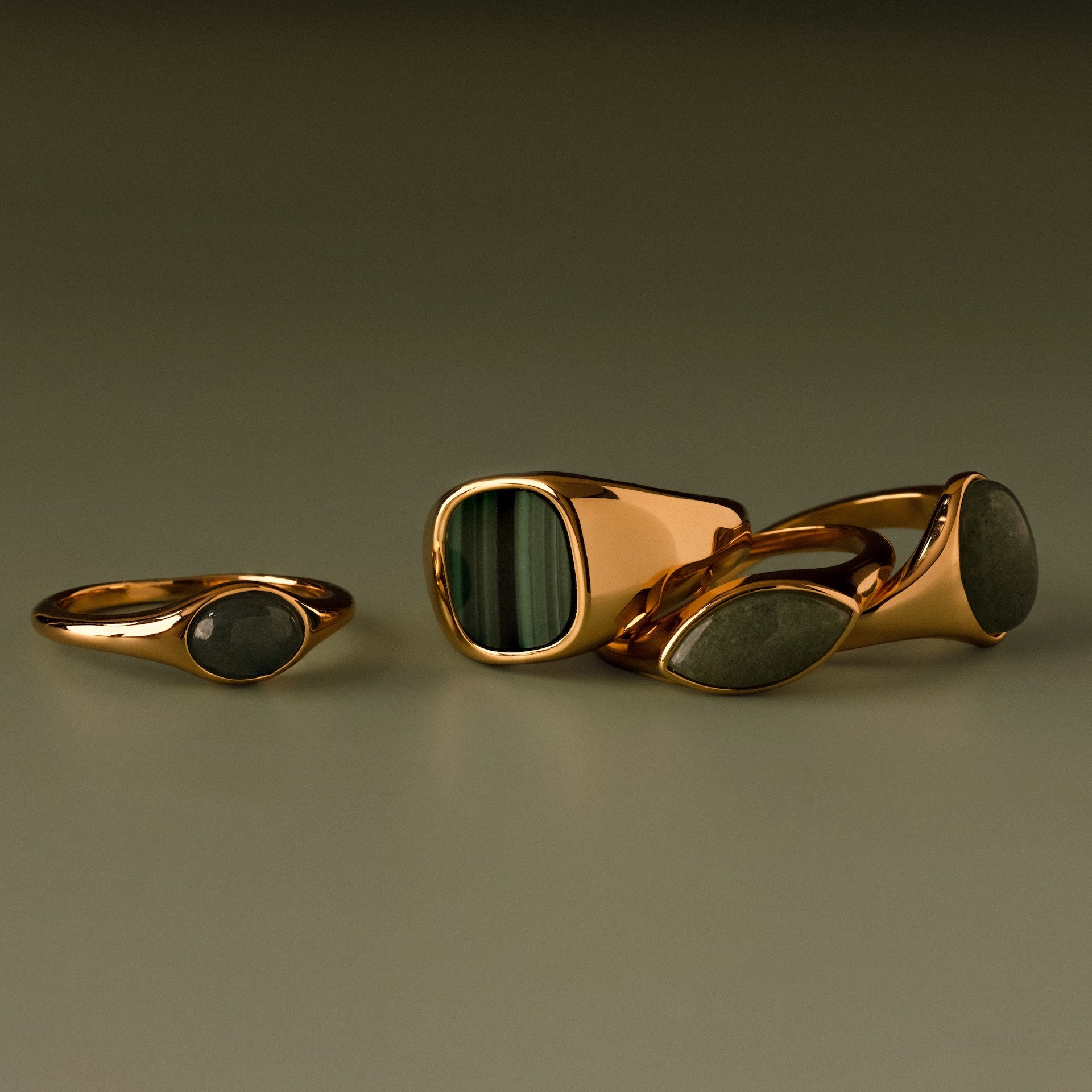 Four gold rings with green and gray stone inlays, including the Malachite Signet Ring—featuring malachite, the stone of transformation—displayed on a neutral surface.